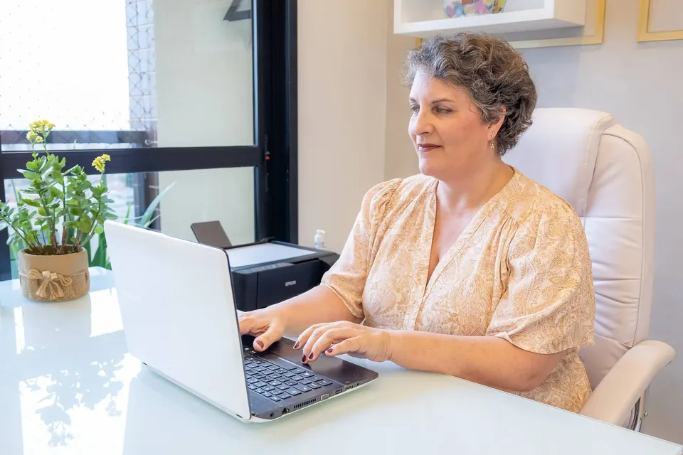 Dra. Patrícia diante do notebook, junto a uma janela com plantas, durante um atendimento online.