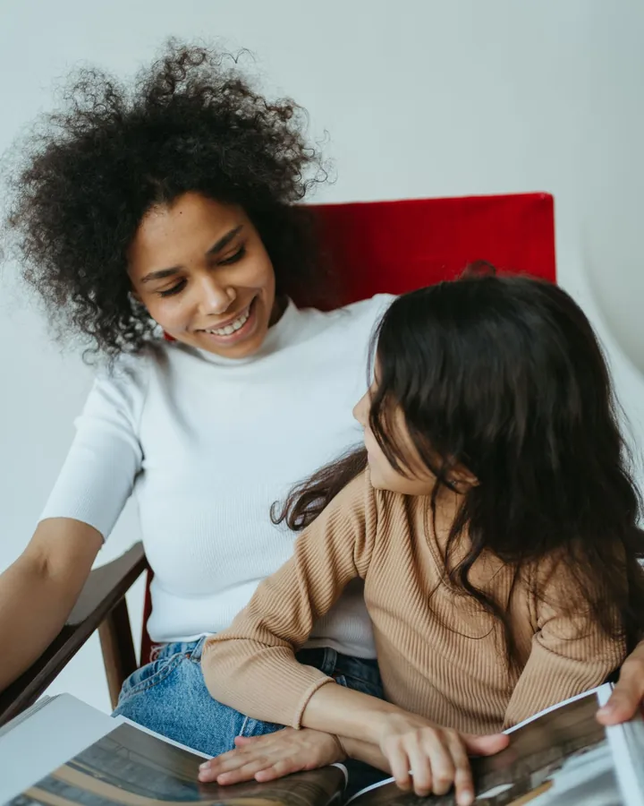 Mãe e filha sentadas juntas numa poltrona, folheando uma revista e sorrindo uma para a outra.